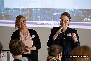 Kurzer Alternativtext: Zwei Frauen sprechen vor einem Publikum mit Mikrofonen in der Hand; im Hintergrund ist eine Leinwand mit einer Projektion zu sehen.
