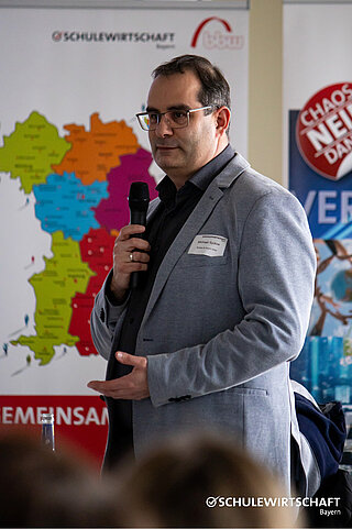 Ein Mann spricht bei einer Veranstaltung vor einer Karte von Bayern und dem Logo von SCHULEWIRTSCHAFT Bayern.
