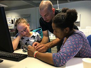 Ein Auszubildender hilft zwei Teilnehmerinnen beim Programmieren. (© Jacqueline Klemm)