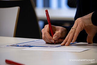 Hände schreiben mit rotem Stift auf Papier, SCHULEWIRTSCHAFT Bayern Logo unten rechts.