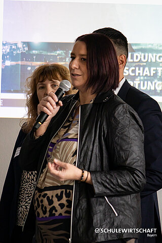  Drei Personen bei einer Präsentation, im Hintergrund ist das Logo "SCHULEWIRTSCHAFT Bayern" sichtbar.