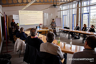 Ein Konferenzraum mit mehreren Personen an einem U-förmigen Tisch, zwei Frauen halten eine Präsentation vor einer Leinwand.