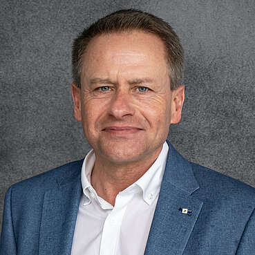 Porträt eines Mannes in Business-Kleidung mit freundlichem Gesichtsausdruck vor grauem Hintergrund.