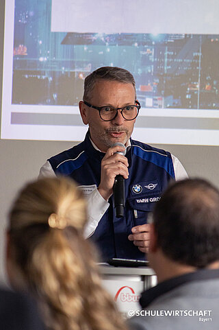 Eine Person hält einen Vortrag an einem Rednerpult, trägt eine BMW-Weste, im Hintergrund ist eine Präsentation zu sehen.