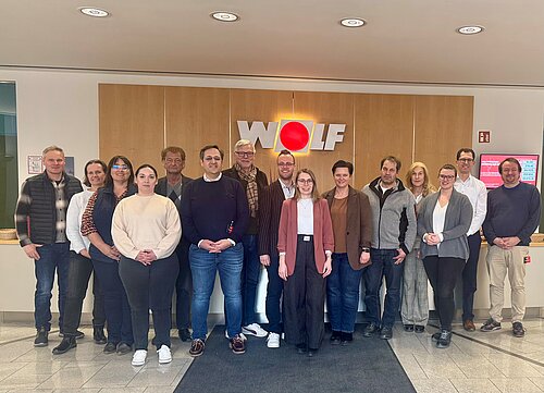 Das Bild zeigt eine Gruppe von 15 Personen, die vor einem Empfangsbereich stehen, auf dessen Wand das Logo "WOLF" prominent von hinten beleuchtet zu sehen ist. Die Gruppe ist in zwei Reihen angeordnet. Im Hintergrund sind Holzverkleidungen.