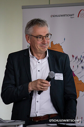 Ein Mann im Anzug hält ein Mikrofon vor einem Banner von SCHULEWIRTSCHAFT Bayern.