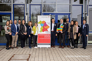 Eine Gruppe von dreizehn Personen steht vor einem Gebäude neben einem Roll-Up-Banner mit dem Text "GEMEINSAM ZUKUNFT GESTALTEN - DAS NETZWERK SCHULEWIRTSCHAFT DEUTSCHLAND".