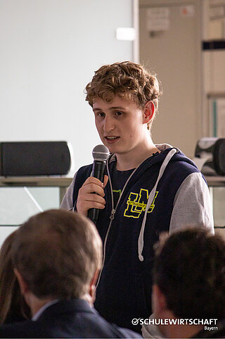 Ein Schüler spricht in ein Mikrofon bei einer Veranstaltung von SCHULEWIRTSCHAFT Bayern.
