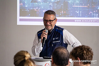 Kurzer Alternativtext: Eine Person spricht in ein Mikrofon an einem Rednerpult, trägt eine BMW Group Weste; im Hintergrund ist eine projizierte Stadtansicht und die URL "www.schulewirtschaft.de" zu sehen.
