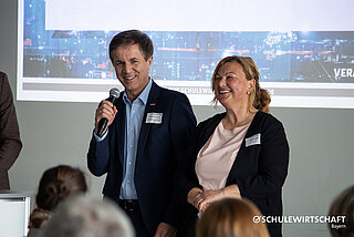 Zwei Personen sprechen vor einem Publikum; eine hält ein Mikrofon.