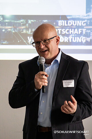 Ein Mann in einem Anzug spricht in ein Mikrofon vor einer Leinwand mit dem Logo von SCHULEWIRTSCHAFT Bayern.