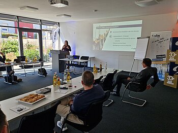 Ein Konferenzraum mit mehreren Personen, die einer Präsentation zuhören. Auf der Leinwand steht "VFM AKADEMIE - EINSTEIGERKURS IT" mit Themen wie IT-Basics, Netzwerke, Datenschutz und Cybercrime. Im Hintergrund sind Banner mit "vfm" und "33 Jahre" zu sehen.