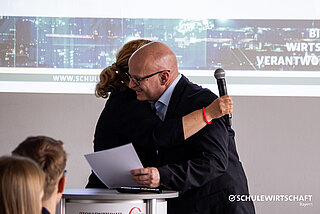 Zwei Personen umarmen sich auf einer Bühne vor einer Präsentationsleinwand.